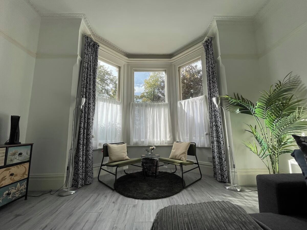 Une pièce d’angle avec une baie vitrée, une table ronde, deux chaises, un lampadaire et une plante en pot.