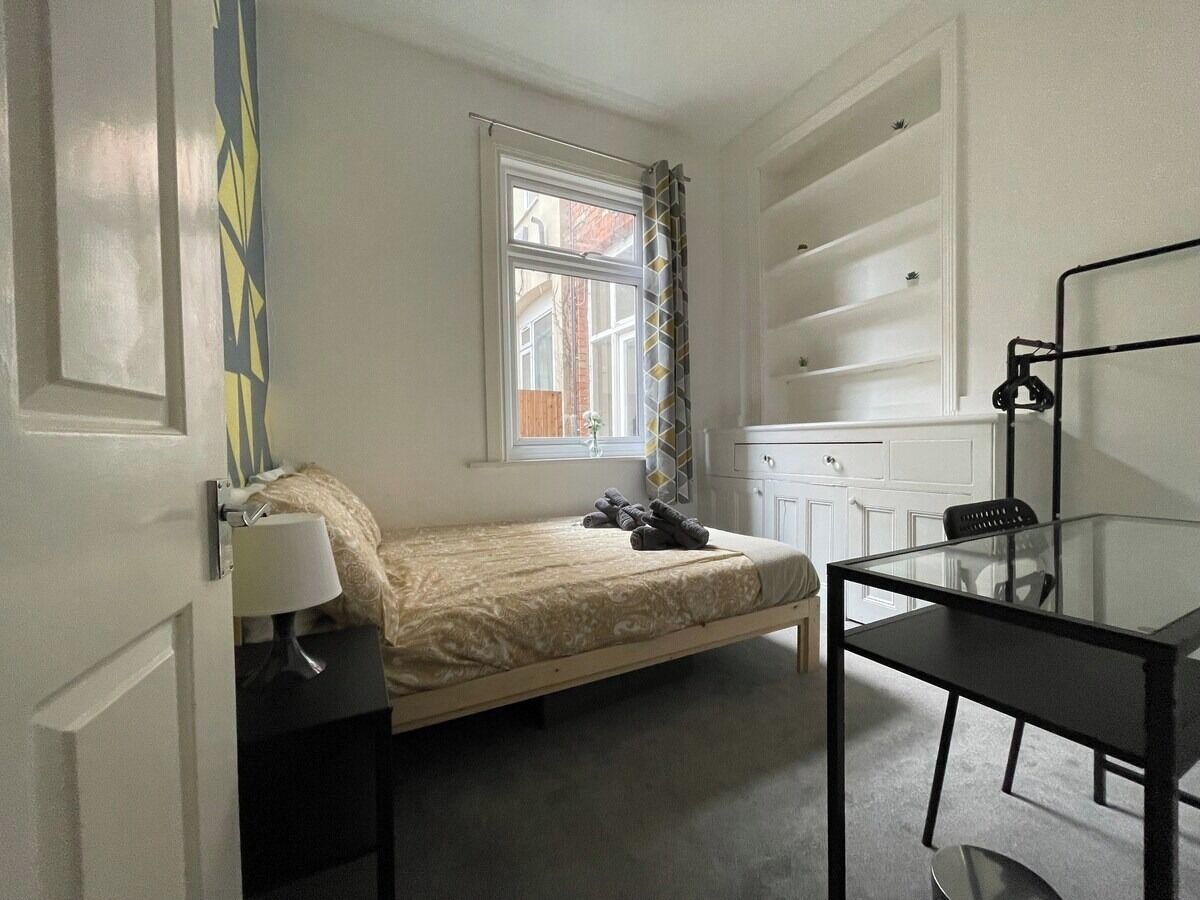 Ein kleines Zimmer mit einem Einzelbett, einem Fenster, einem Nachttisch mit Lampe, einem Glastisch und einem Stuhl.