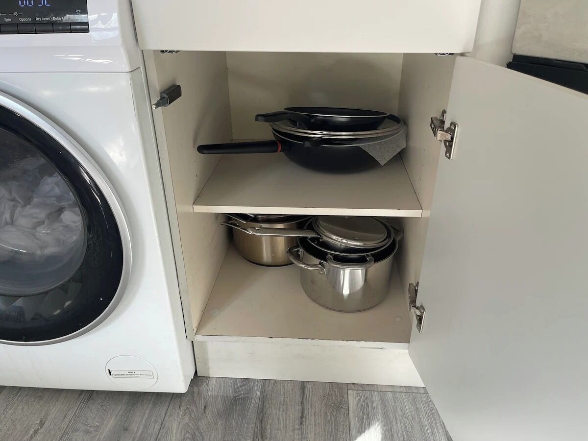 Una cocina con lavadora, una sartén negra y dos ollas de acero inoxidable en estantes.