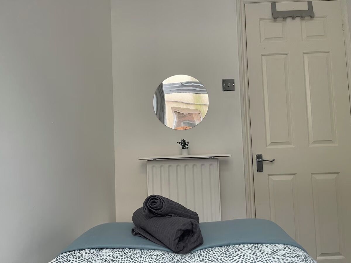 A bedroom with a bed, a folded towel, a radiator, a mirror, and a door.