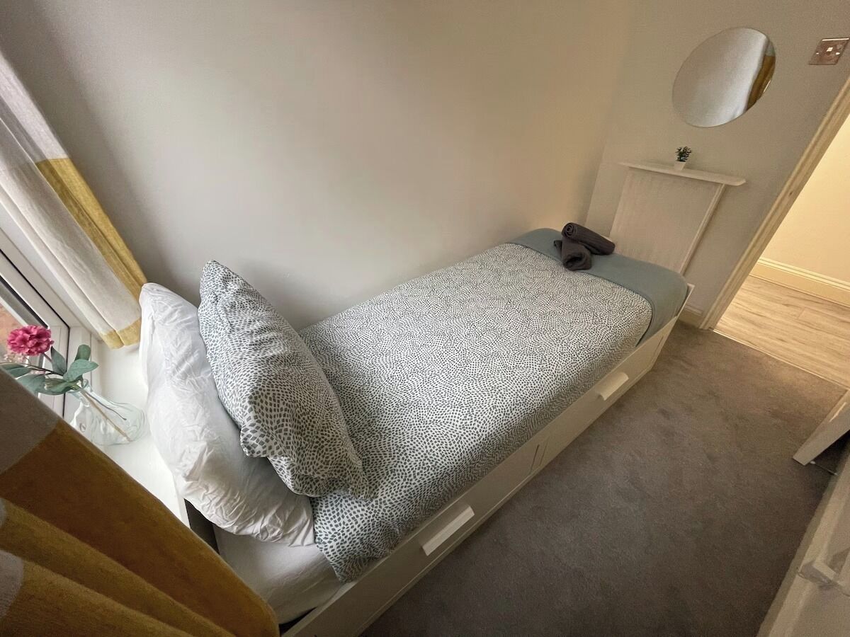 Une petite chambre avec un lit simple, recouverte d’une courtepointe à motifs blancs et gris, un oreiller blanc et une table de chevet avec un miroir.
