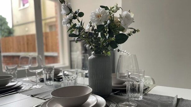 Un tavolo da pranzo apparecchiato con piatti, ciotole, posate e un vaso con fiori bianchi.