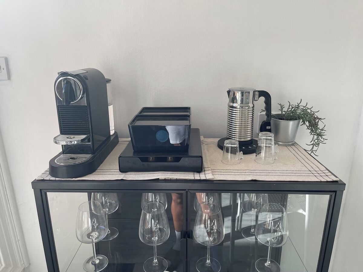 Een koffiezetapparaat, een waterkoker en een kleine plant in pot op een tafel met daaronder een glazen kast met wijnglazen.