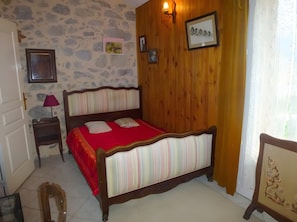 A bedroom with a wooden bed, a stone wall, a wooden door, a nightstand, a lamp, and a framed picture on the wall. - Farm Cottage "La Grange" (Garde-Colombe)