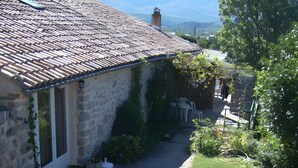 A stone building with a tiled roof, a small garden with white plastic chairs and a table, and a view of distant mountains. - Farm Cottage "La Grange" (Garde-Colombe)