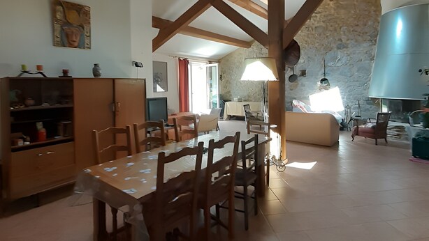 A spacious kitchen with a dining area, wooden beams, a stone wall, and a large metal water heater. - Farm Cottage "La Grange" (Garde-Colombe)
