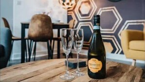 Une bouteille de vin Prosecco et deux verres sur une table en bois, dans un intérieur moderne agrémenté de motifs hexagonaux sur les murs et d’une horloge.