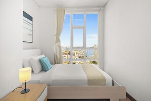 Ein modernes Hotelzimmer mit großem Fenster, einem Bett mit weißer Bettwäsche, einem Nachttisch mit Lampe und Blick auf die Stadtlandschaft.