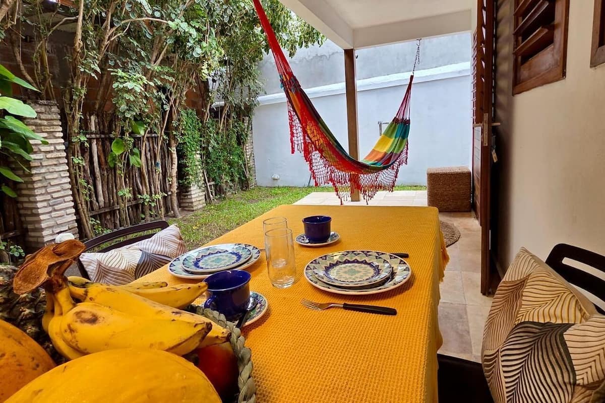A dining area with a table set for two, a hammock, and a view of a garden with trees and a white wall.