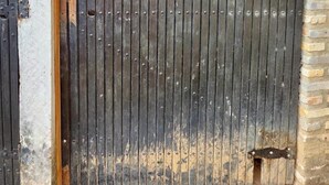 Una verja de metal con barras verticales y un cerrojo, adosada a un muro de ladrillo.