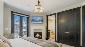 A bedroom with a fireplace, a large bed, a TV, and a balcony with a view of houses. - Tranquil Blackstone Getaway 3 Bedroom Townhouse (Park City)