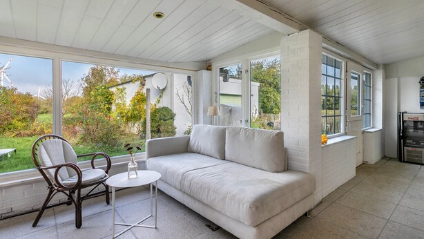 A room with a white sofa, a wicker chair, a small table, and large windows offering a view of greenery and a wind turbine.