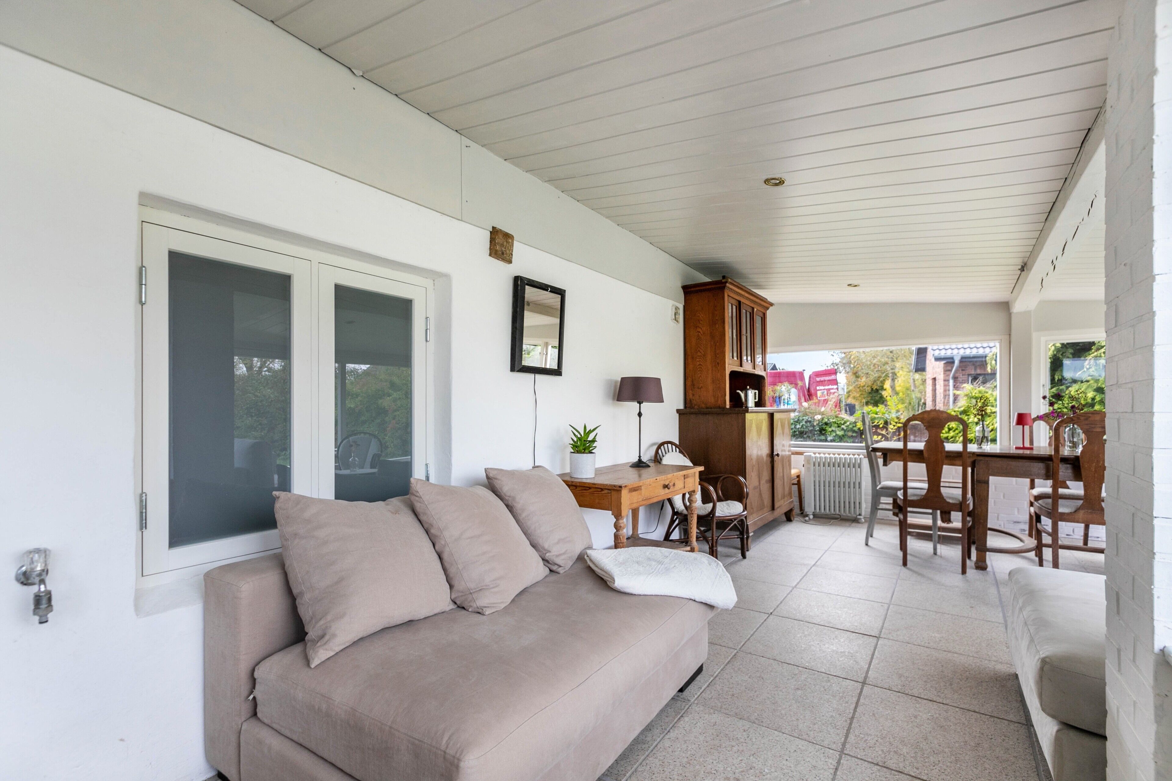 Ein überdachter Patio-Bereich mit einem beigefarbenen Sofa, einem Holzesstisch, Stühlen und einem Schrank.