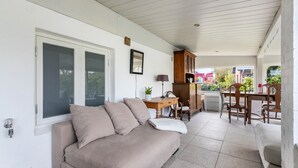 A covered patio area with a beige sofa, wooden dining table, chairs, and a cabinet.