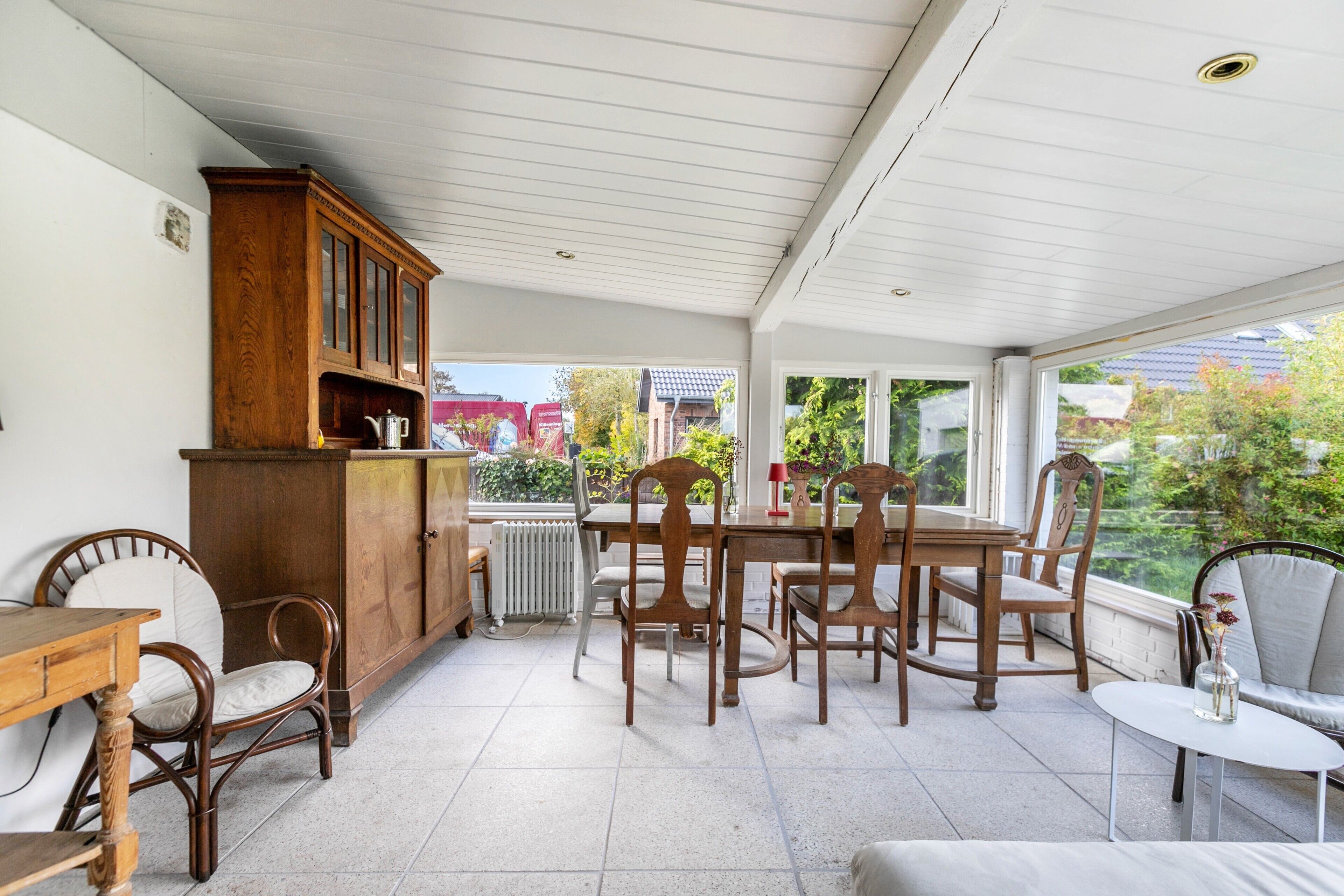 Eine überdachte Terrasse mit Esstisch, Stühlen, einem Schrank und einem kleinen Tisch mit einer Vase.