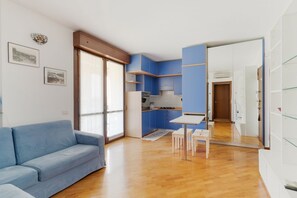 A modern living room with a blue sofa, a dining table with chairs, a kitchen with blue cabinets, and a large mirror reflecting the space.