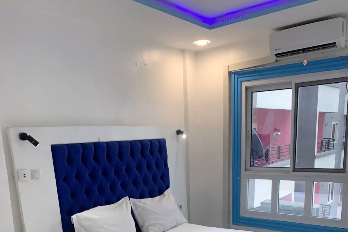 A bedroom with a bed, a bedside table, a window, and an air conditioning unit.