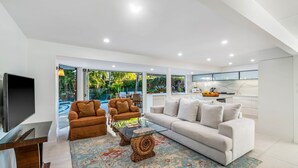 A modern living room with a white sofa, brown armchairs, and a glass coffee table. There is a TV on the left and a kitchen area with a fruit bowl on the counter.