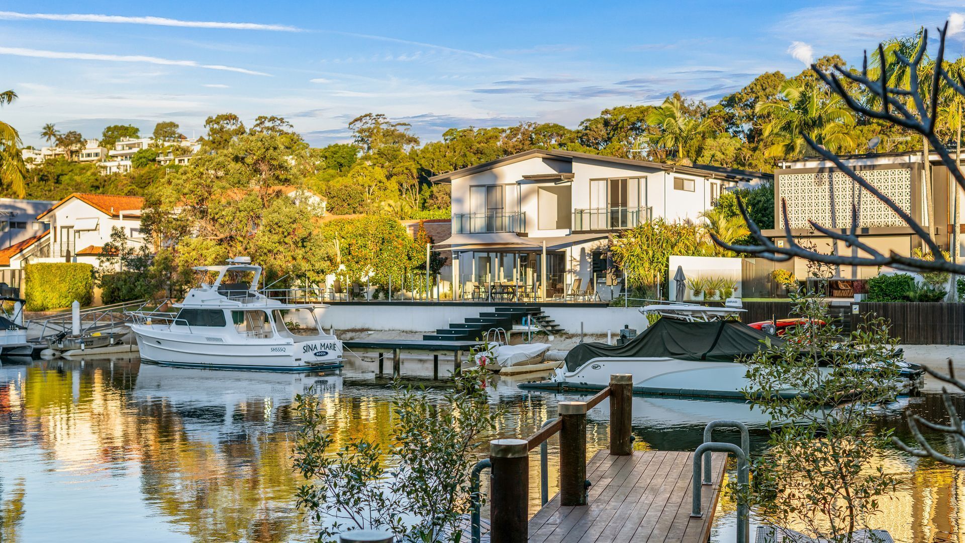 A marina with boats docked, a modern house with a balcony overlooking the water, and a wooden deck in the foreground.