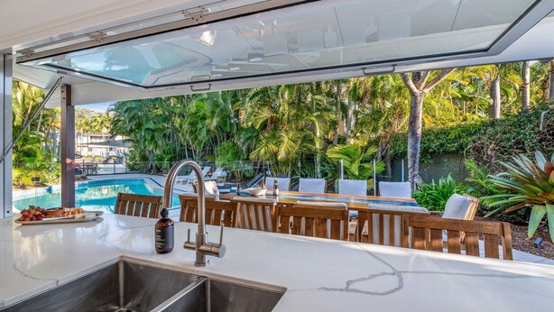 A modern outdoor kitchen with a pool view, wooden furniture, and a glass enclosure.