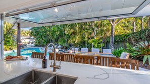 Eine moderne Außenküche mit Blick auf einen Pool, Holzmöbel und einer Glaseinfriedung.