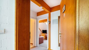 Um corredor com vigas de madeira, uma parede de tijolos brancos e uma porta que dá para um quarto com uma televisão e um sofá de couro marrom.
