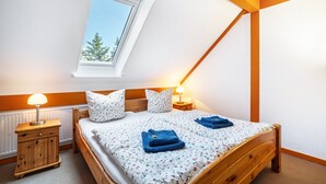 Quarto com teto inclinado, janela, cama com roupa de cama floral, duas luminárias de cabeceira e uma mesa de cabeceira de madeira.