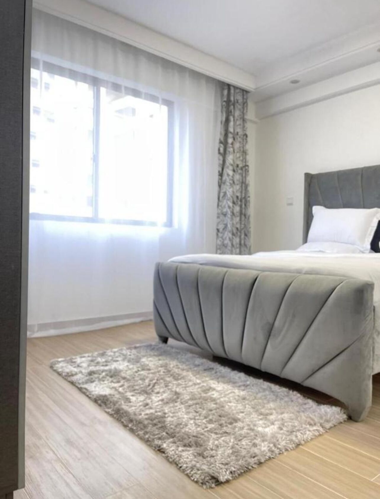 A bedroom with a bed, a window with curtains, a rug, and a wooden floor.