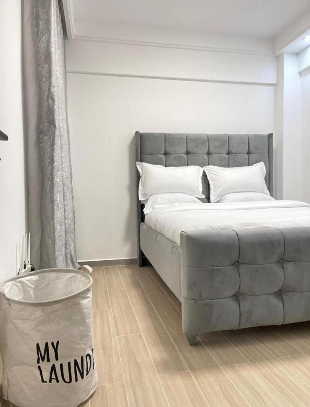 A bedroom with a grey tufted headboard, white bedding, and a grey curtain.