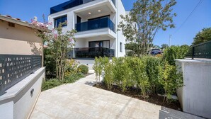 Une maison moderne à deux étages, dotée d’un balcon, entourée d’un jardin où poussent différentes espèces de plantes et agrémentée d’une allée pavée.