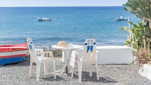 A seaside terrace with two white plastic chairs, a small round table, and a red and blue boat on the pebbled shore. - Holiday Home 'Callao 1' with Sea View, Wi-Fi and Air Conditioning (Playa Quemada)