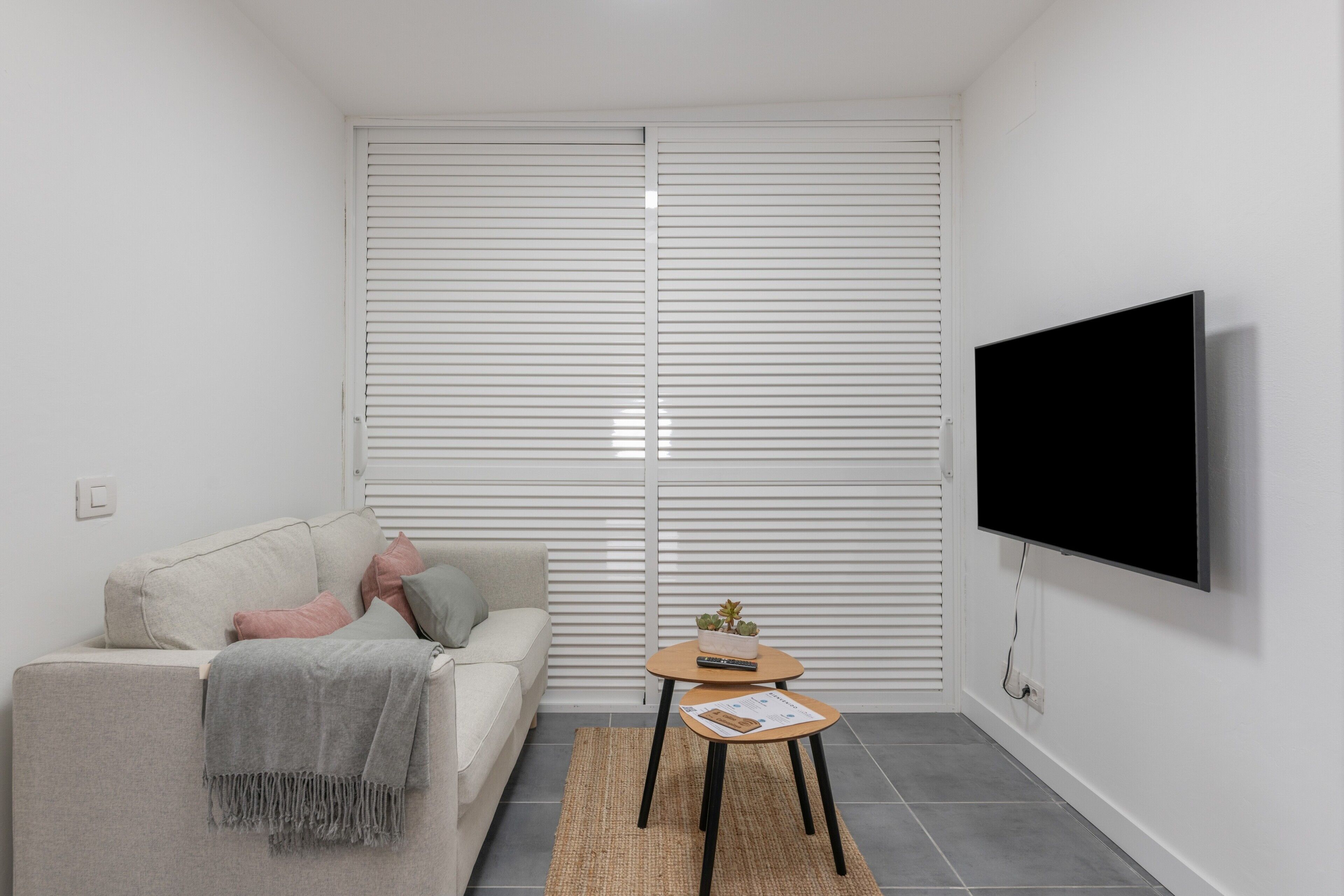 Ein modernes Wohnzimmer mit einer Couch, einem kleinen Tisch, einem Fernseher und einem Fenster mit Jalousien.