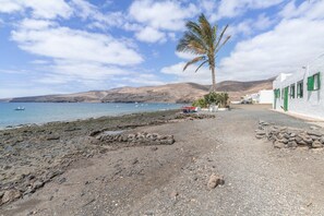 A coastal scene with a rocky beach, a palm tree, and white buildings with green shutters. - Casa Vacacional 'Callao 1' con Vista al Mar, Wi-Fi y Aire Acondicionado (Playa Quemada)