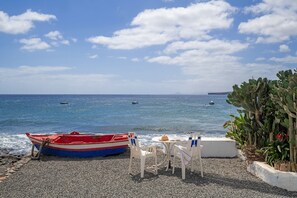 A pebbled beach with a red and blue boat, two white plastic chairs, and a table with a hat. - Holiday Home 'Callao 2' with Sea View, Wi-Fi and Air Conditioning (Playa Quemada)