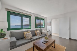 Una sala de estar moderna con un sofá gris, una mesa de centro de madera y vistas al mar a través de amplios ventanales.