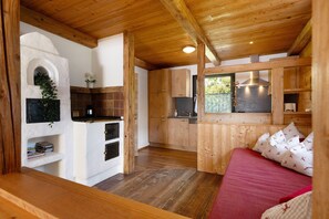 L’intérieur chaleureux d’un chalet avec un plafond en bois, une cheminée en pierre, un coin cuisine et un lit recouvert d’une literie rouge.