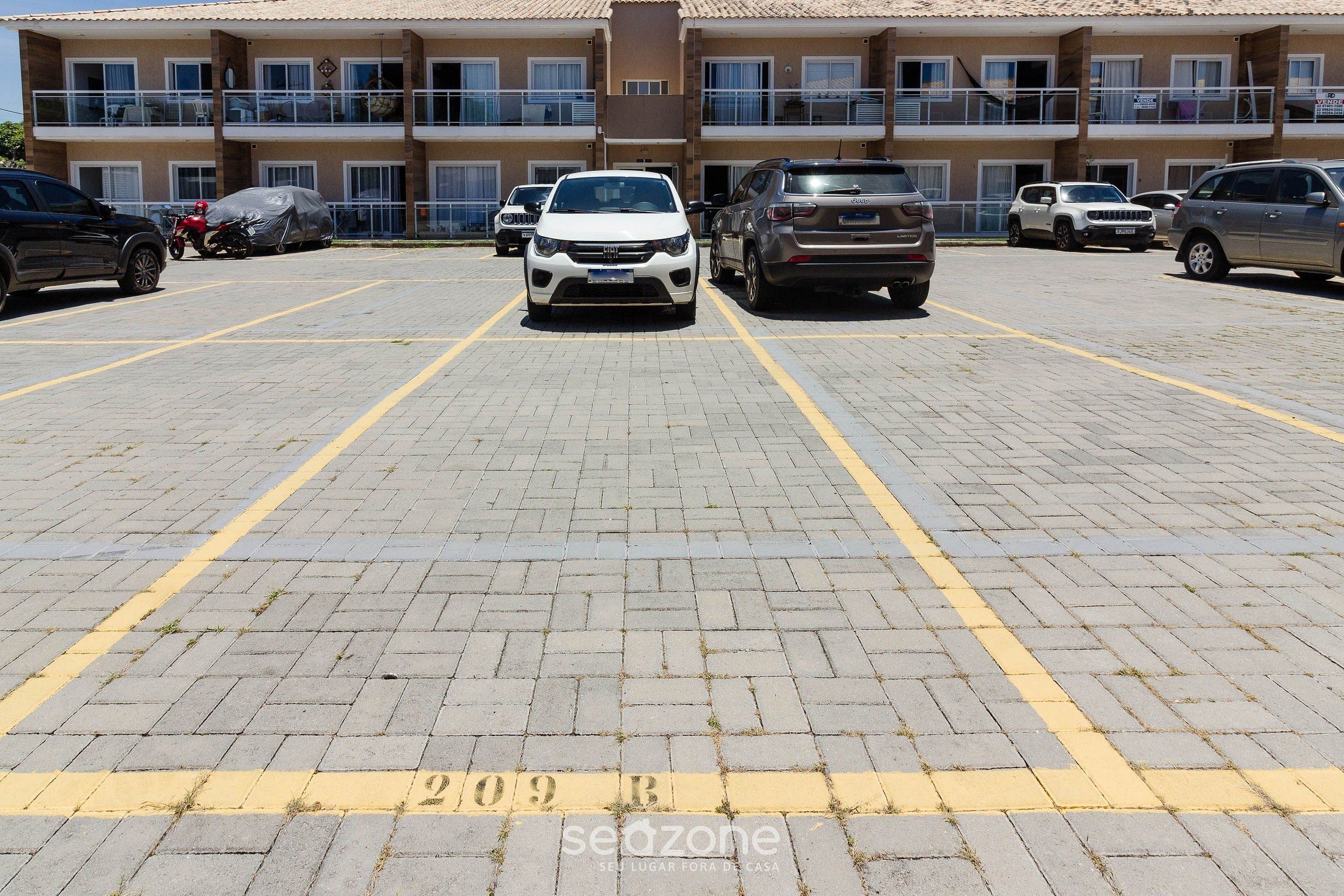Un parcheggio con diverse auto e uno scooter, un edificio con balconi e un terreno asfaltato con linee gialle.