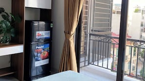 Habitación de hotel con cama, minibar, planta en maceta, puerta corrediza a un balcón y vistas de los edificios exteriores.