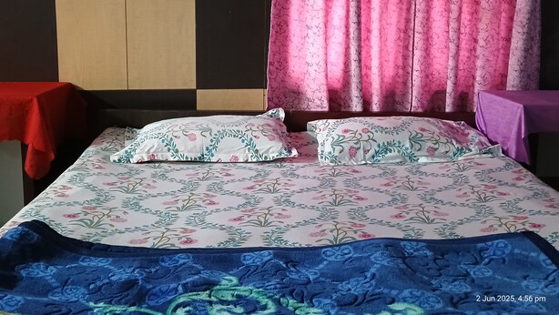 A bed with floral bedding, a red table on one side, and a purple table on the other. - Swaroop Ashray (Siliguri)