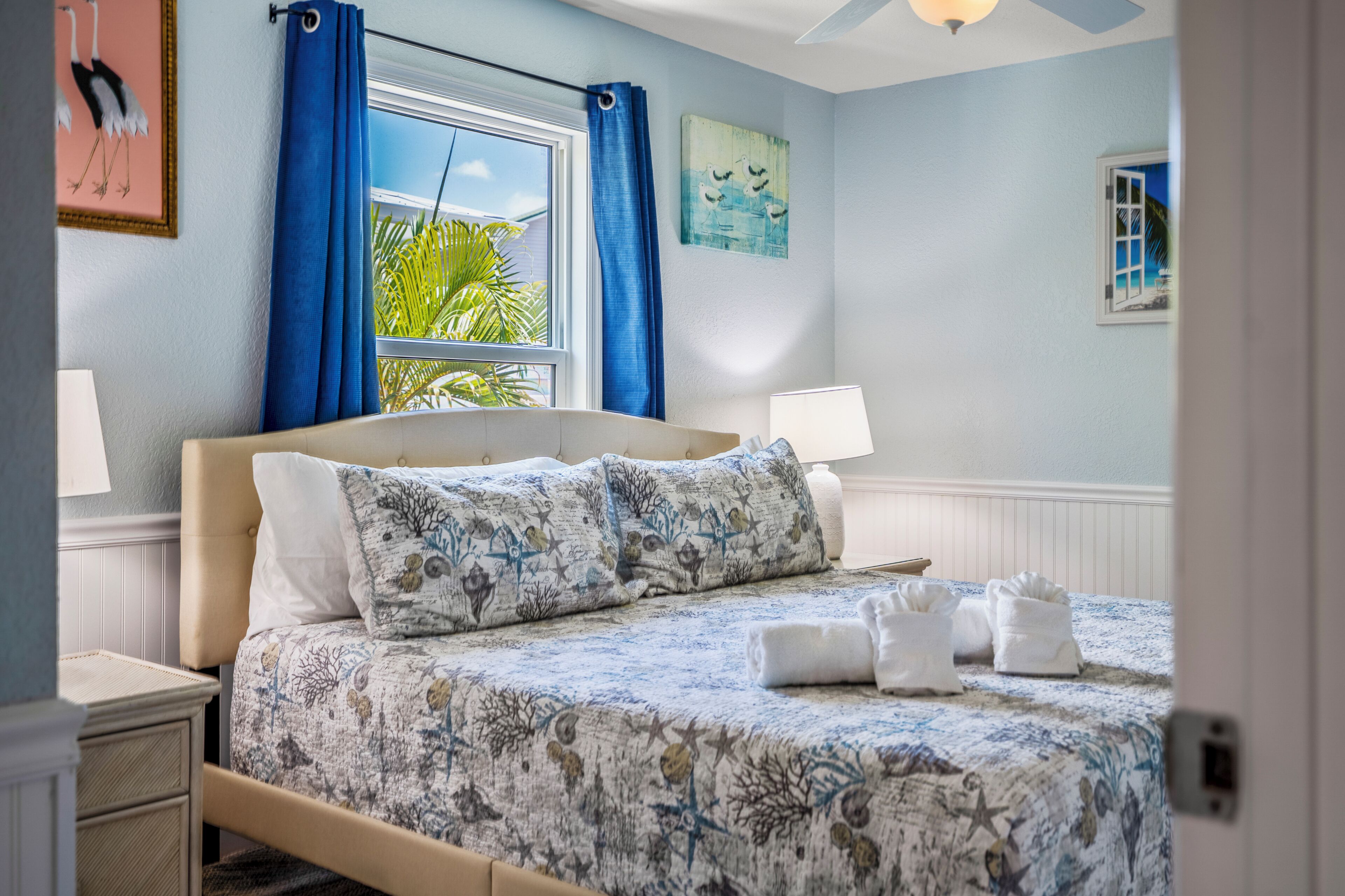 Een slaapkamer met een bed, een raam met gordijnen, een nachtkastje, een lamp en kunstwerken aan de muur.