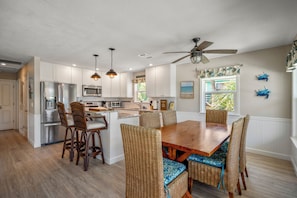 A kitchen with white cabinets, stainless steel appliances, wicker chairs, and a wooden dining table. - Ostego Charm: Brand new pool, porch, lush landscape—walk-able to the Beach! (Fort Myers Beach)