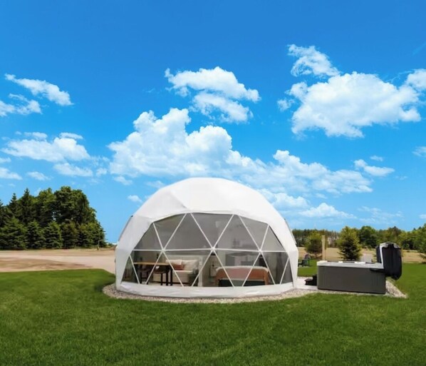 A geodesic dome structure with a transparent roof, surrounded by a grassy field and trees, under a clear blue sky. - The Sunset Dome- a private escape, with breathtaking sunsets, rooted in nature. (Guelph/Eramosa)
