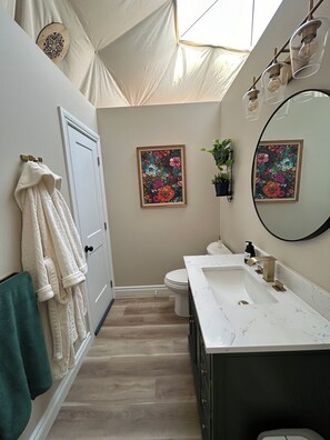 A bathroom with a white sink, dark cabinet, and a large mirror. There are two framed floral paintings on the wall, a towel rack with robes, and a door with a decorative panel. - The Sunset Dome- a private escape, with breathtaking sunsets, rooted in nature. (Guelph/Eramosa)