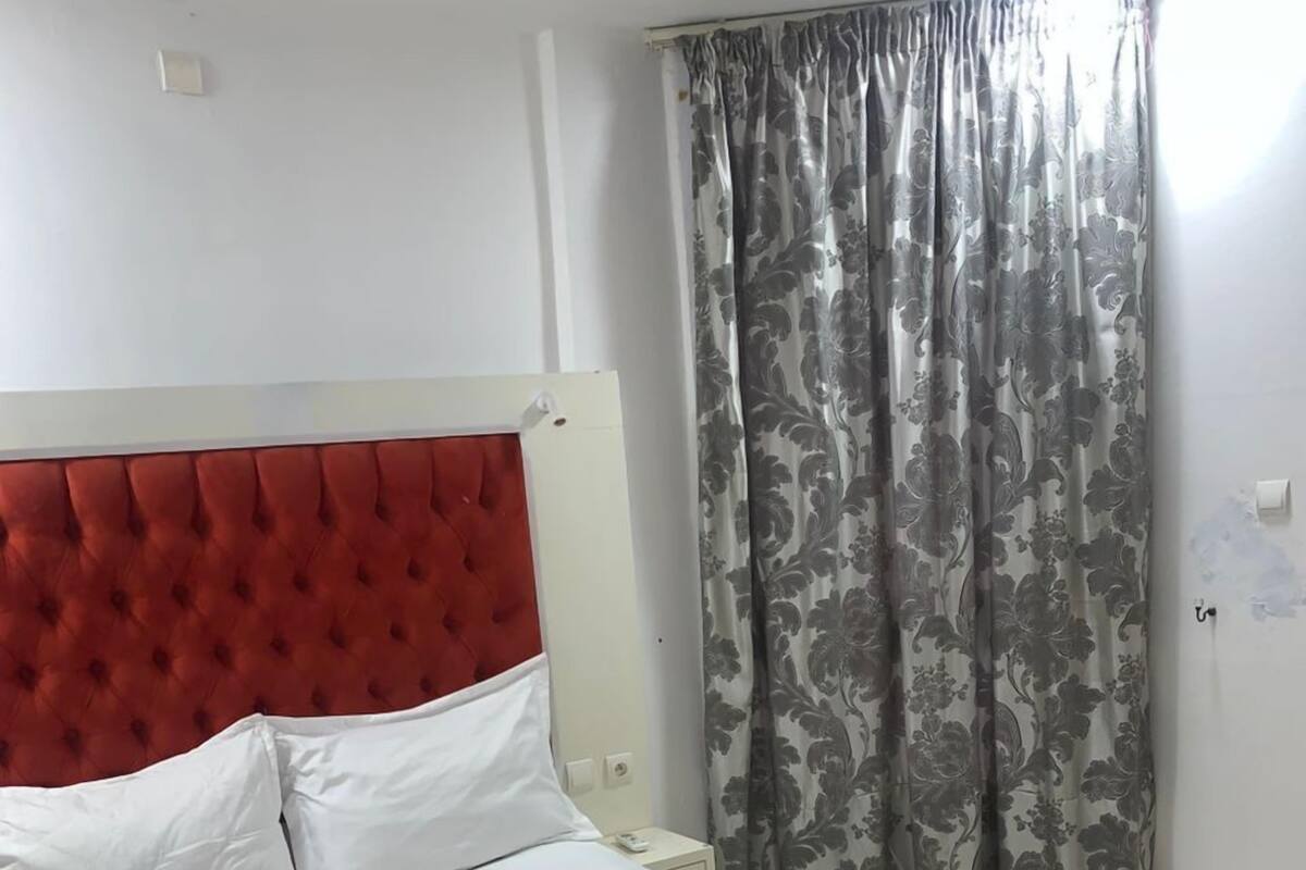 A bed with a red headboard and white bedding, a small bedside table, and a window with floral curtains.