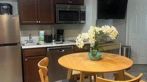 A modern kitchen with dark wooden cabinets, a stainless steel refrigerator, a microwave, and a coffee maker. There is a round wooden table with two chairs and a vase of flowers. - Condo Near Disney No Resort Fee Free Parking (Kissimmee)