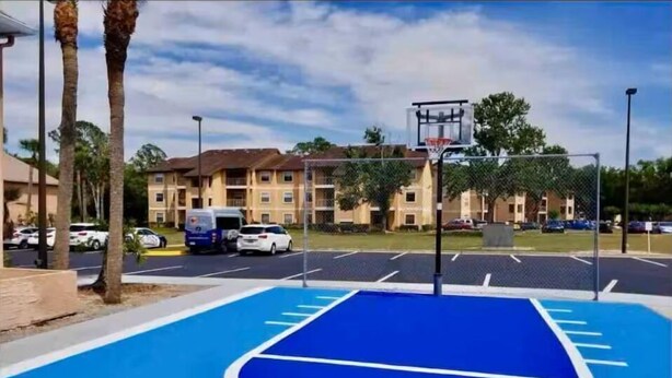 A basketball court with a blue surface and white lines, surrounded by a parking lot with cars and a building in the background. - Condo Near Disney No Resort Fee Free Parking (Kissimmee)