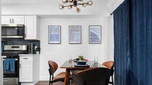 Une cuisine moderne avec des armoires blanches, un four à micro-ondes en acier inoxydable et un four encastré. On y trouve une table à manger avec quatre chaises et un rideau bleu sur le côté droit.