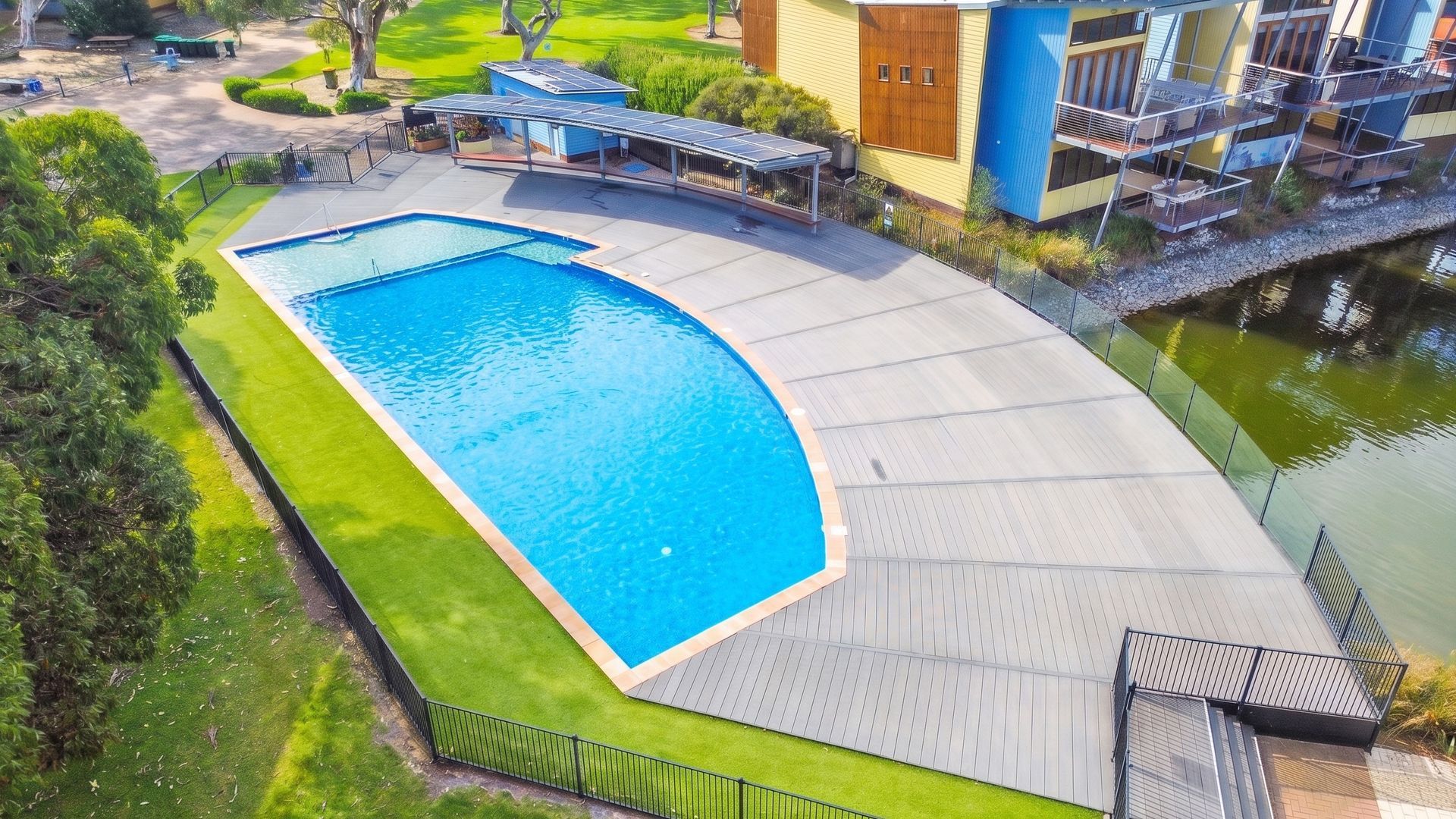 A swimming pool with a curved deck, adjacent to a building with a blue facade and a balcony.