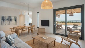 A modern living room with a sofa, coffee table, and dining area. There is a large window with a view of the outdoors. - Near Naousa |3BR Villa| Private Pool |Villa Candia (Paros)