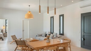 A modern dining area with a wooden table and chairs, a woven pendant light, and a sofa with cushions. - Near Naousa |3BR Villa| Private Pool |Villa Candia (Paros)